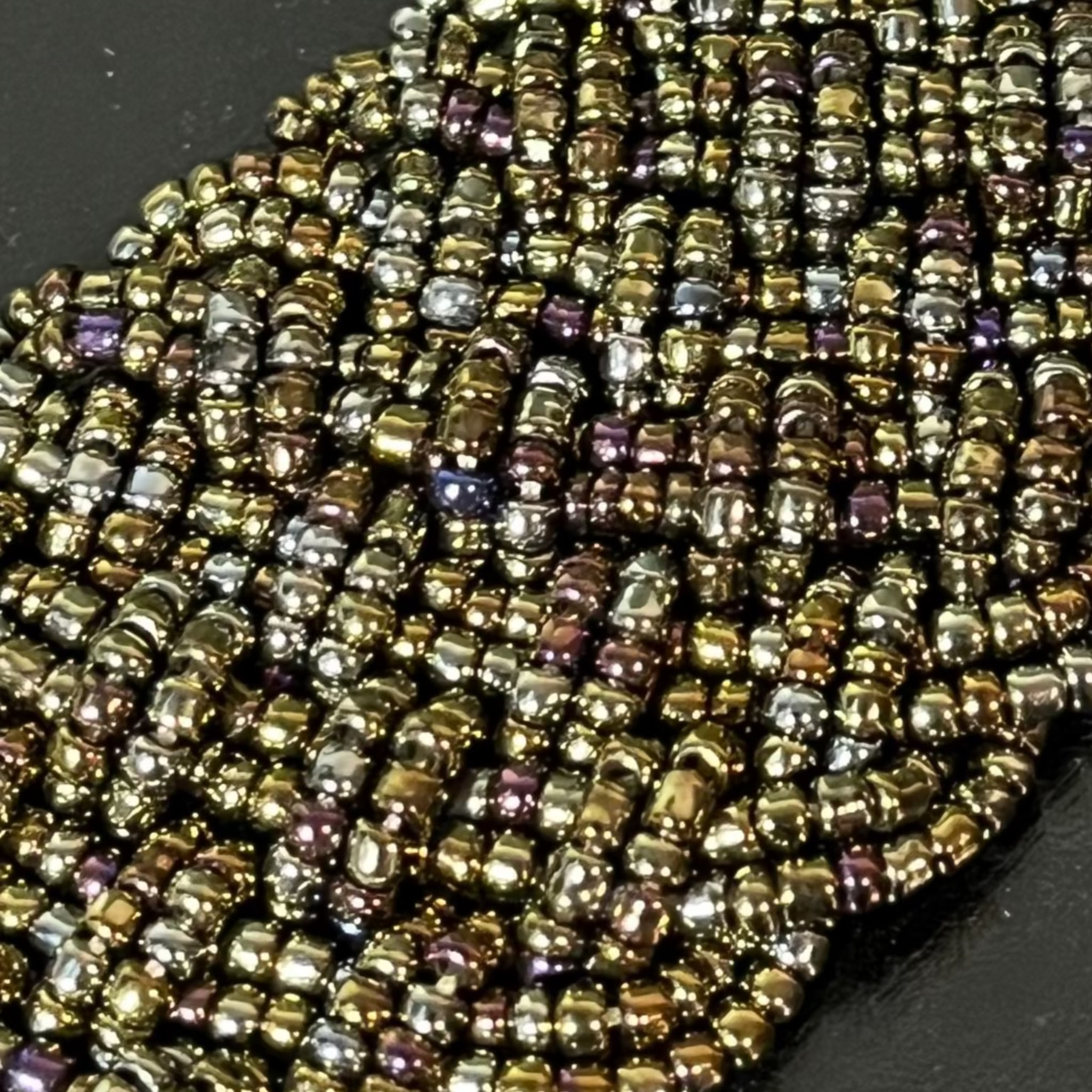 A close-up of a handmade beaded collar or trim made of small metallic beads in a wavy pattern, featuring gold, bronze, and iridescent purple accents on a dark background.