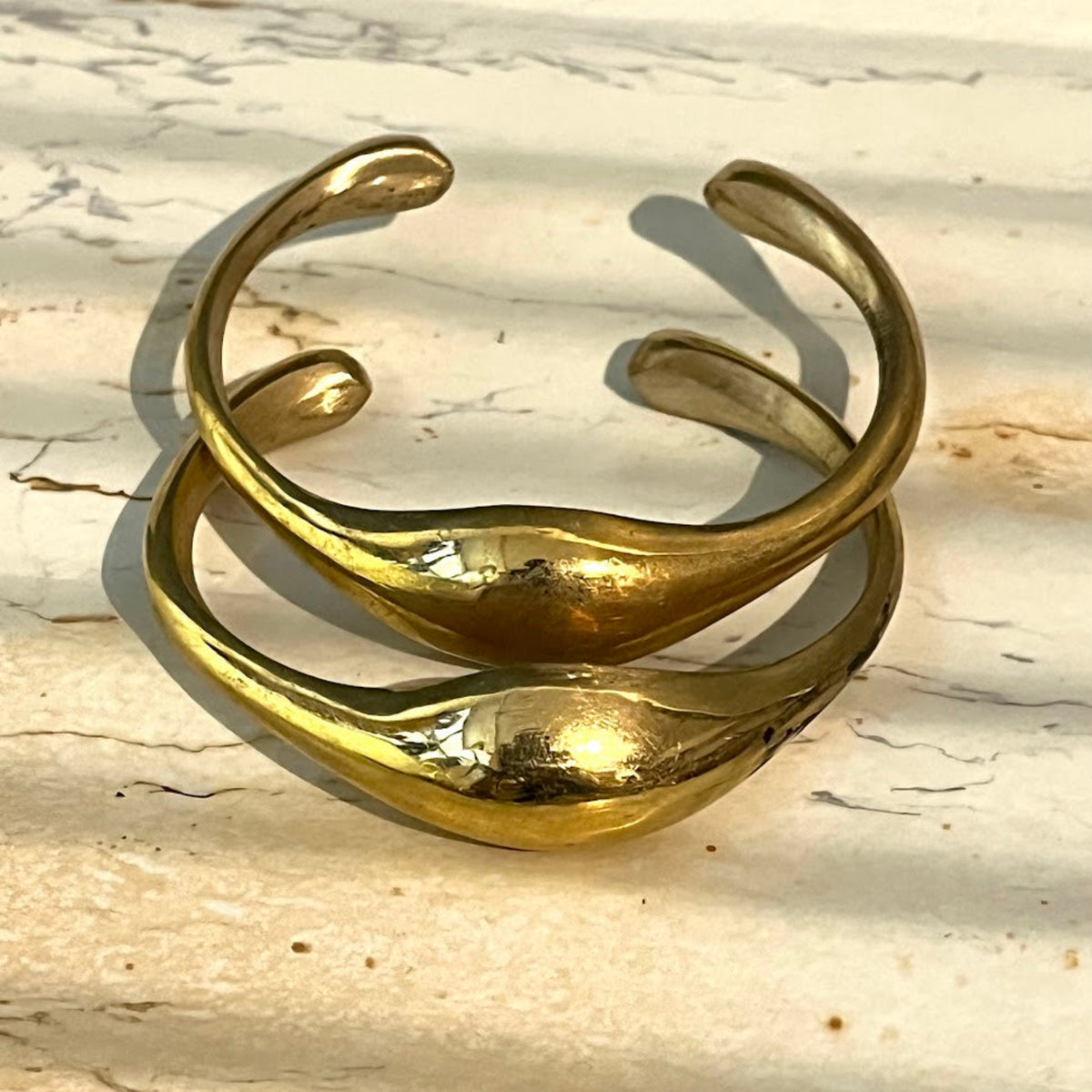 A gold-toned open cuff bracelet with a smooth, organic wavy design, photographed on a light beige stone surface with natural shadows.