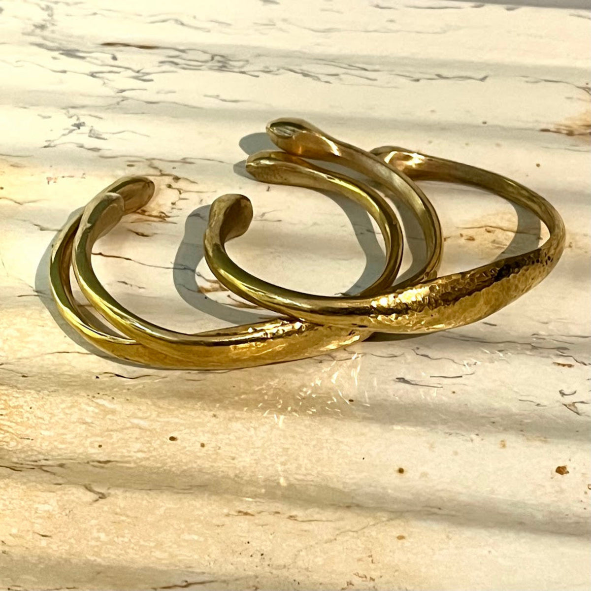 Three hammered gold-tone open cuff bracelets arranged on a light beige stone surface with natural shadows.