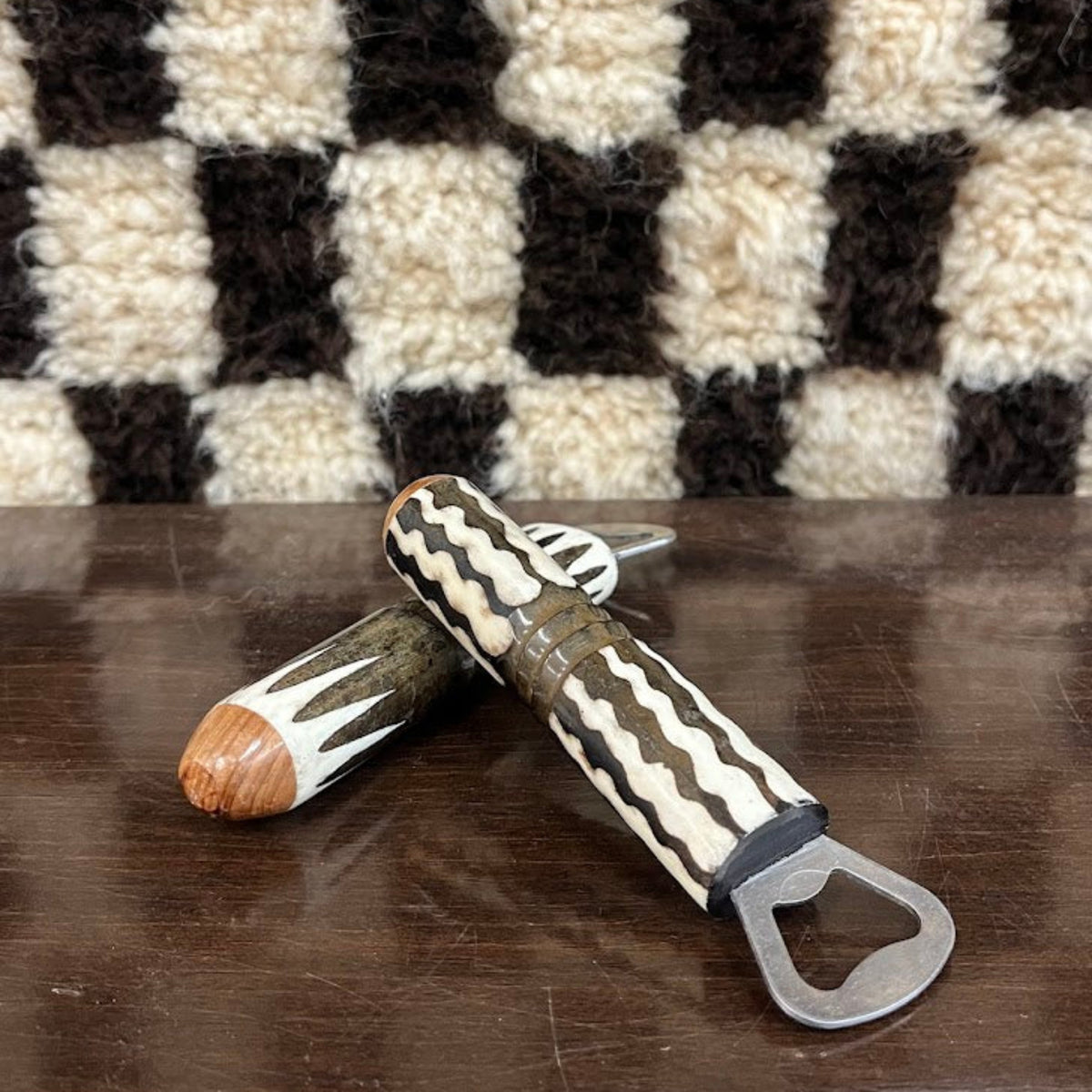 Two bottle openers with carved wooden handles and metal pull tabs, resting on a dark wooden surface in front of a black and cream checkered fuzzy rug.