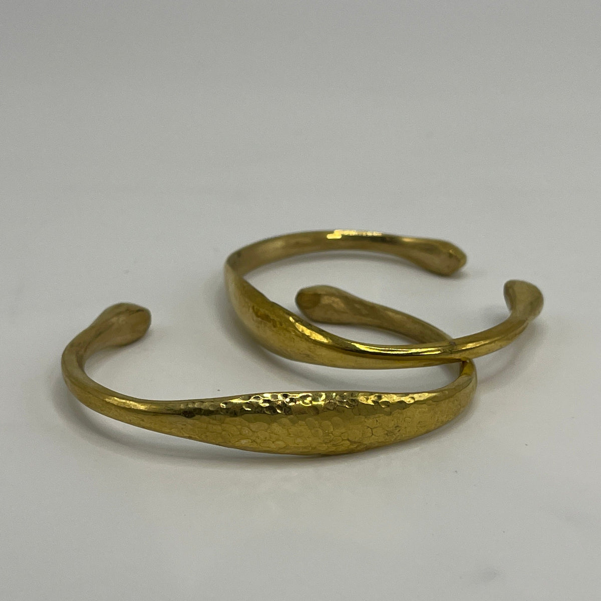 A textured gold-tone open cuff bracelet with a hammered finish, displayed on a neutral light gray background.