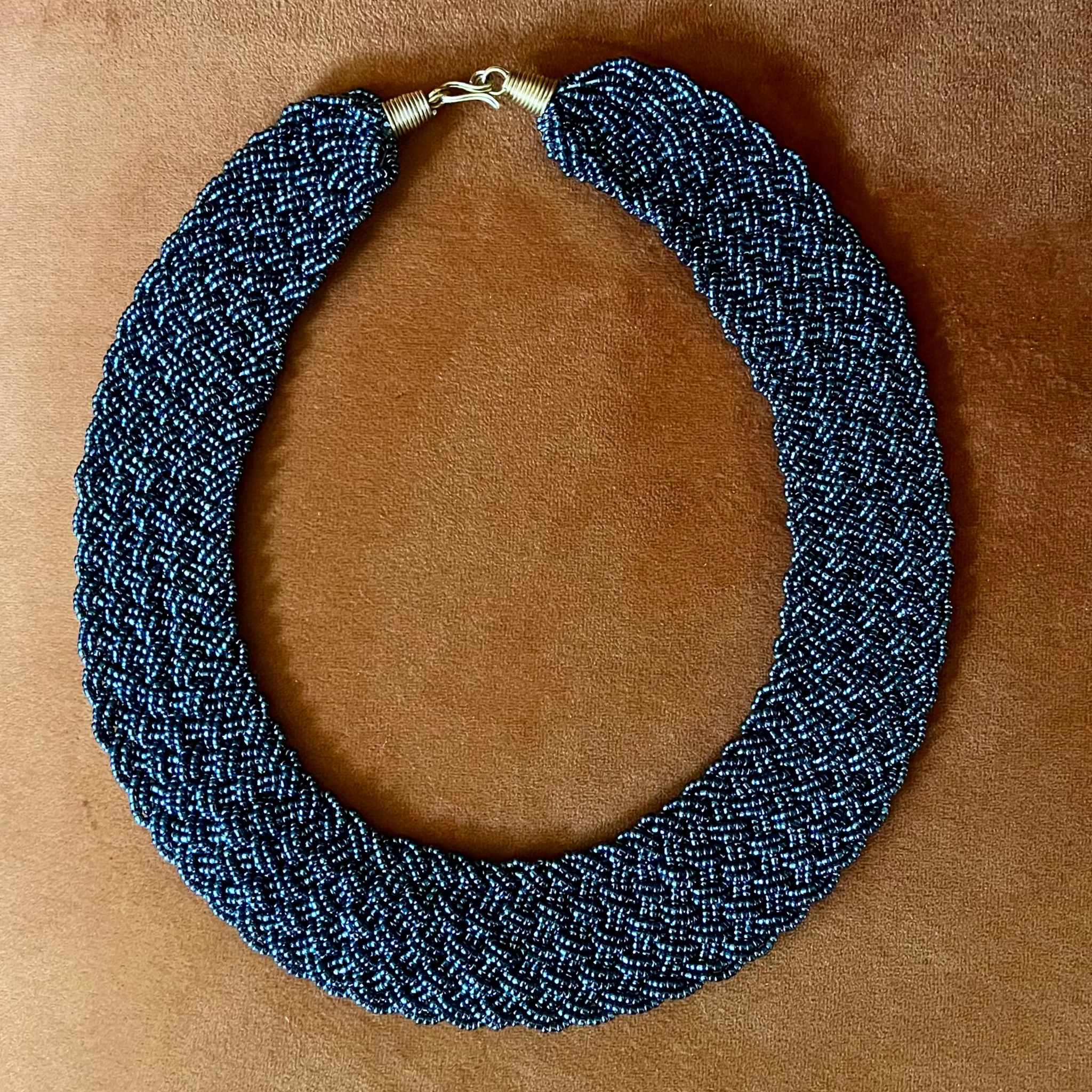 A thick braided woven collar necklace in a dark blue and black pattern, featuring a small gold clasp, laid flat on a brown suede-like background.